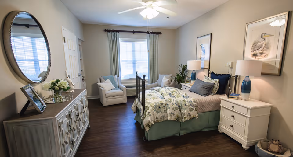 A cozy bedroom with a bed featuring floral bedding and multiple pillows, two white nightstands with blue lamps, framed bird artwork on the walls, a comfortable armchair near a window with curtains, a decorative mirror above a sideboard with flowers and a photo frame, and wooden flooring.