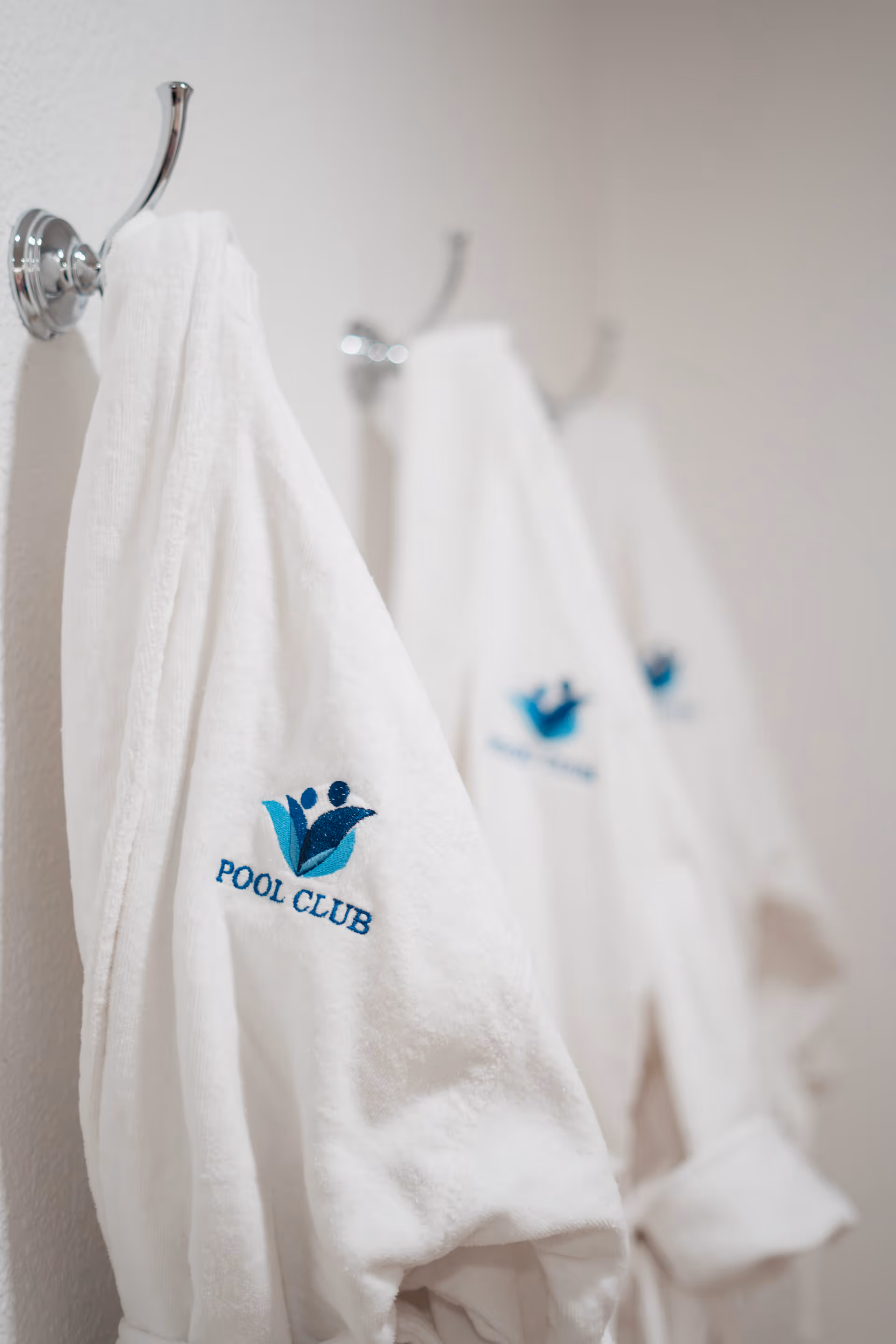 Three white bathrobes hanging on chrome hooks on a white wall, each embroidered with a blue and teal logo and the words 'POOL CLUB'.