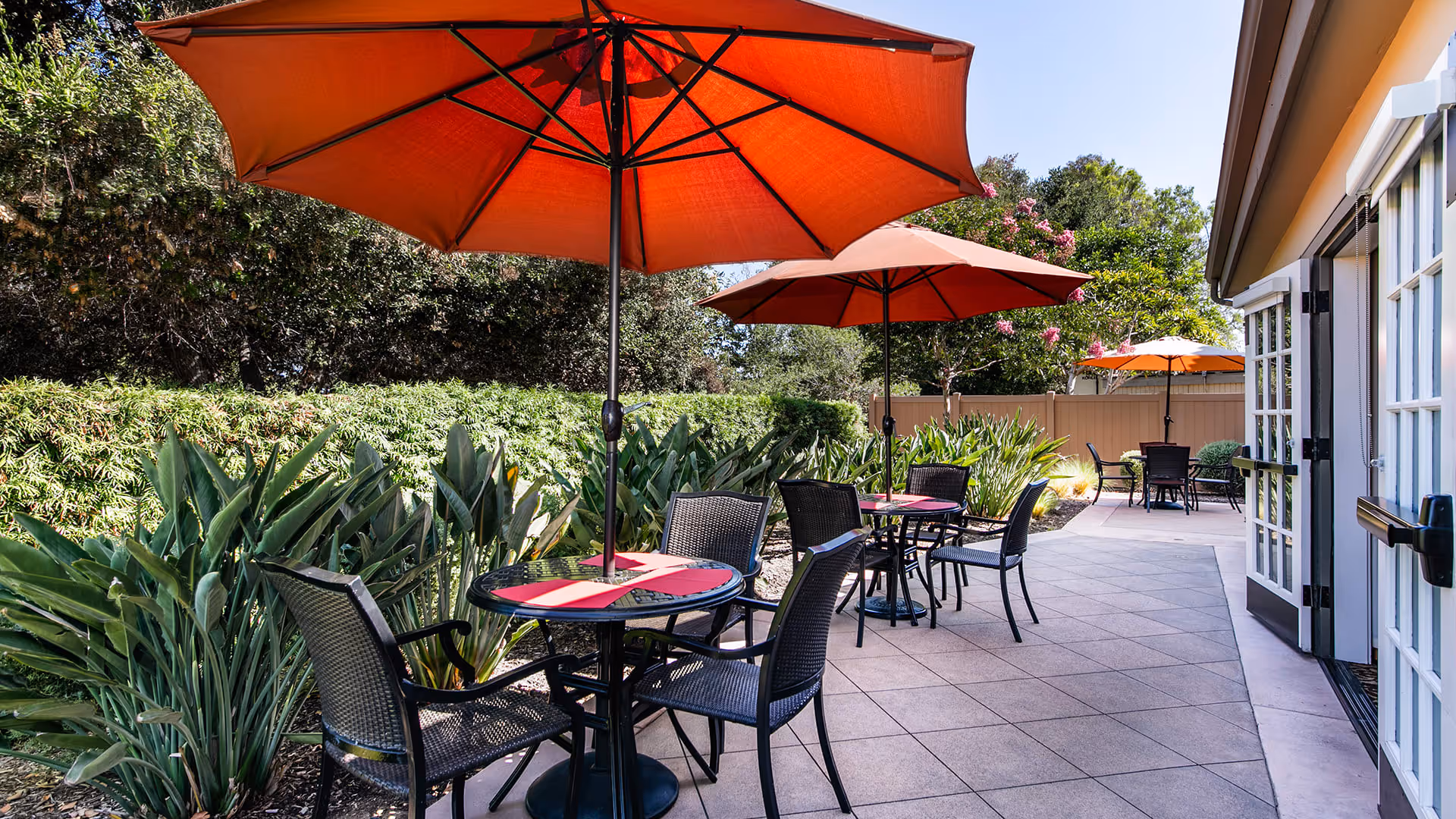 Outdoor patio area with several round tables, each with four black wicker chairs and large red umbrellas providing shade. The patio is surrounded by green plants and trees, and there is a building with white-framed glass doors on the right side.