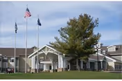 Exterior view of the New York State Veterans Home building with a large tree in front and three flagpoles displaying the American flag and other flags on a grassy lawn under a partly cloudy sky.