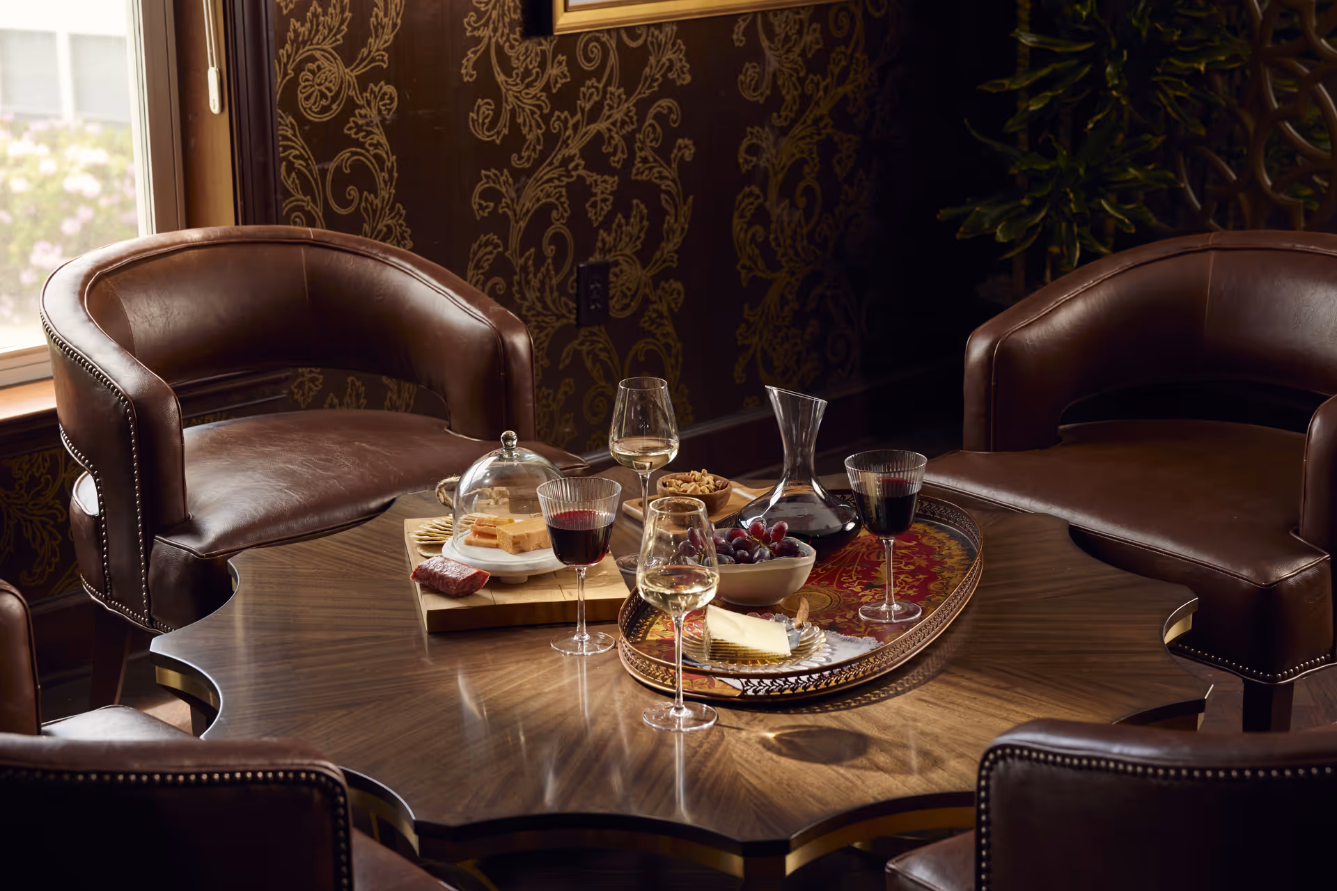 A cozy seating area with four brown leather chairs around a wooden table. On the table, there is a decorative tray with a bowl of grapes, a glass decanter, and several glasses of red and white wine. There is also a wooden board with cheese, crackers, and nuts. The background features a window with a view of flowers outside and a wall with ornate patterned wallpaper.