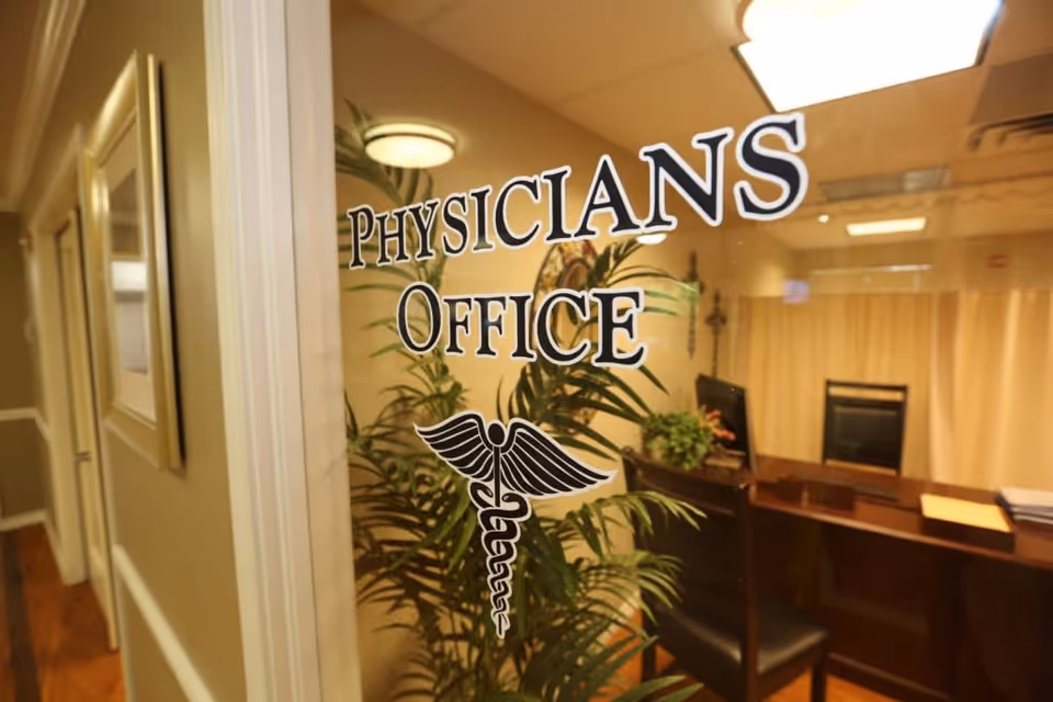 View through a glass door labeled 'Physicians Office' with a caduceus symbol, showing a reception desk, chair, and indoor plants inside a medical office area.