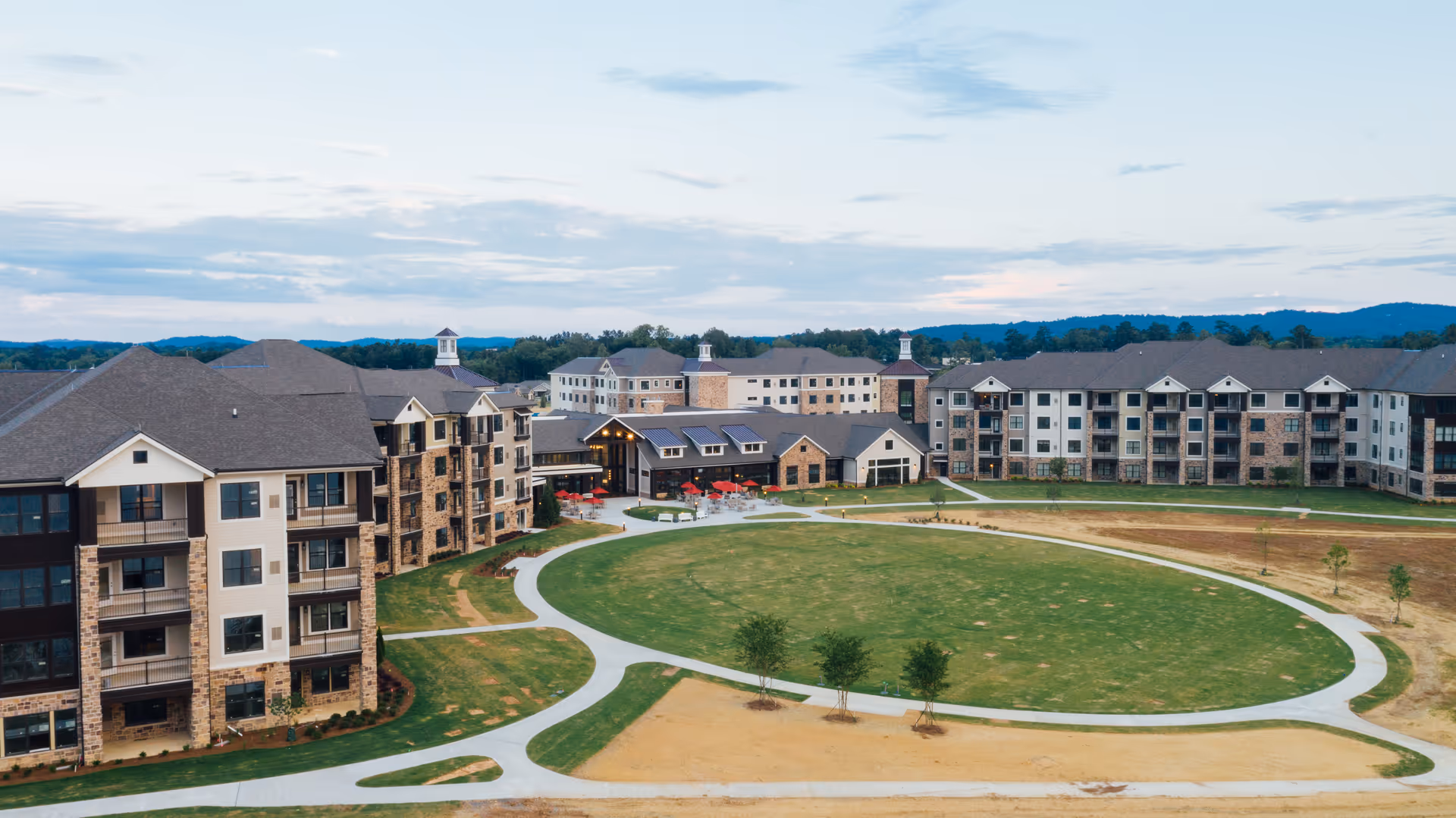 Aerial view of Health Services at The Spires senior living facility showing multiple connected buildings surrounding a large oval-shaped grassy courtyard with walking paths and small trees, set against a backdrop of distant hills under a partly cloudy sky.