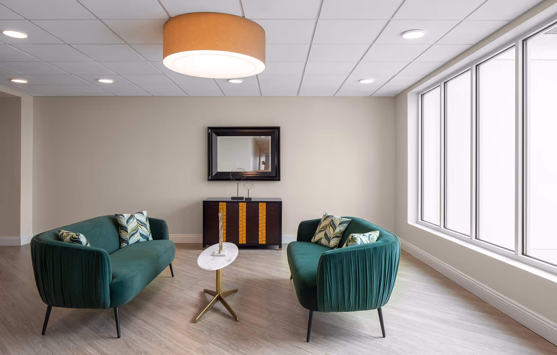 A modern sitting area with two green upholstered sofas facing each other, each with patterned cushions. Between them is a small oval marble coffee table with a decorative item. Behind the table is a dark wooden cabinet with a mirror hanging above it. The room has large windows on the right side allowing natural light to fill the space, light-colored walls, and a ceiling with recessed lighting and a large round pendant light.