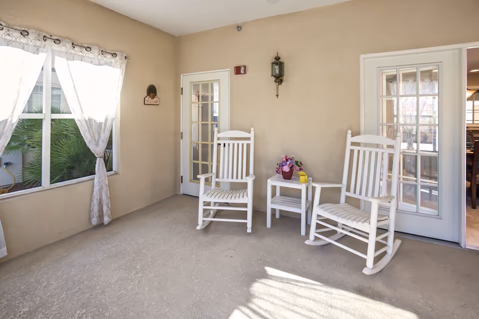 Sunlit enclosed sitting area with two white rocking chairs, a small side table with flowers, and French doors.