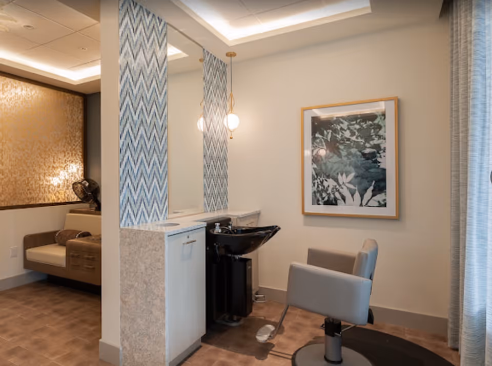 Interior view of a salon area in an assisted living facility featuring a hair washing station with a black sink and a gray salon chair. The wall behind the sink has a large mirror with blue and white chevron patterned panels on either side. A framed artwork with a botanical theme hangs on the adjacent wall. To the left, there is a seating area with a cushioned bench and a decorative gold-patterned wall.