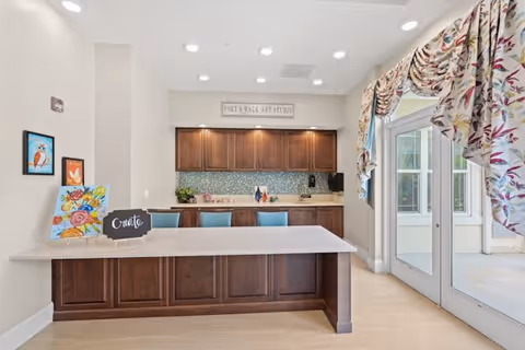 A bright art studio space with a large countertop island in the center, wooden cabinets along the back wall, and a mosaic tile backsplash. The wall above the cabinets has a sign that reads 'Poet's Walk Art Studio'. There are colorful paintings and art supplies on the countertop and walls. To the right, there are glass doors with floral curtains leading to an outdoor area.