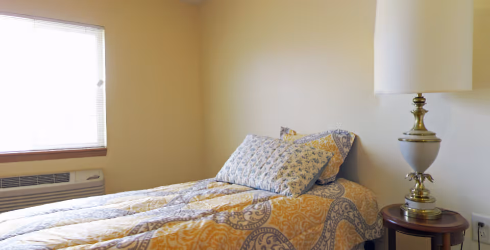 Small furnished bedroom with a patterned twin bed, decorative pillows, a side table lamp, and a window.