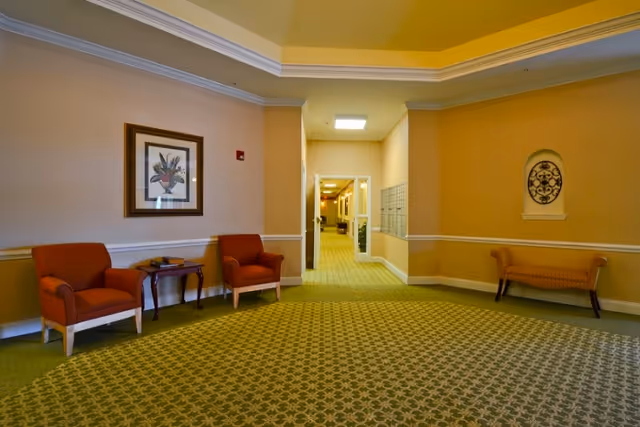 A hallway area in a senior living facility with patterned green carpet, beige walls with white trim, two orange armchairs with a small wooden table between them on the left, a framed floral artwork above the chairs, a bench on the right side, and a corridor leading to other rooms in the background.