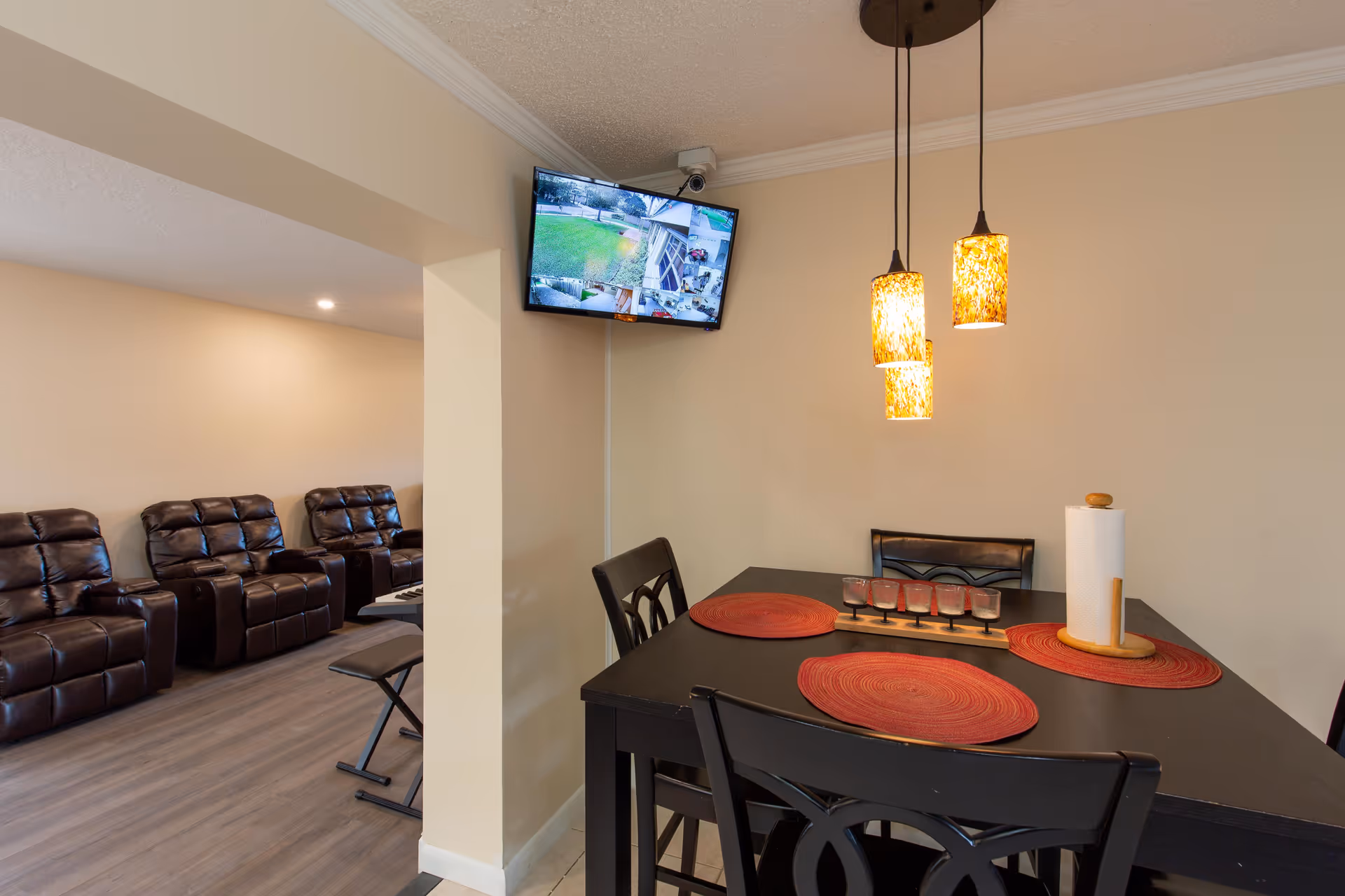 Interior view of a dining area with a black table set with red placemats, a paper towel holder, and a decorative candle holder. Three pendant lights with amber-colored shades hang above the table. Adjacent to the dining area is a living room with brown leather recliners and a keyboard on a stand. A wall-mounted TV screen displays multiple security camera feeds.