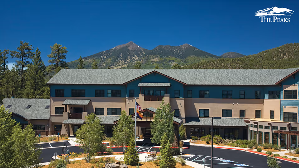Exterior view of The Peaks, A Senior Living Community building with a mountainous backdrop under a clear blue sky. The building is three stories tall with a combination of beige and blue exterior walls, surrounded by trees and landscaped greenery. There is a circular driveway with parking spaces and a flagpole with flags in front of the entrance.