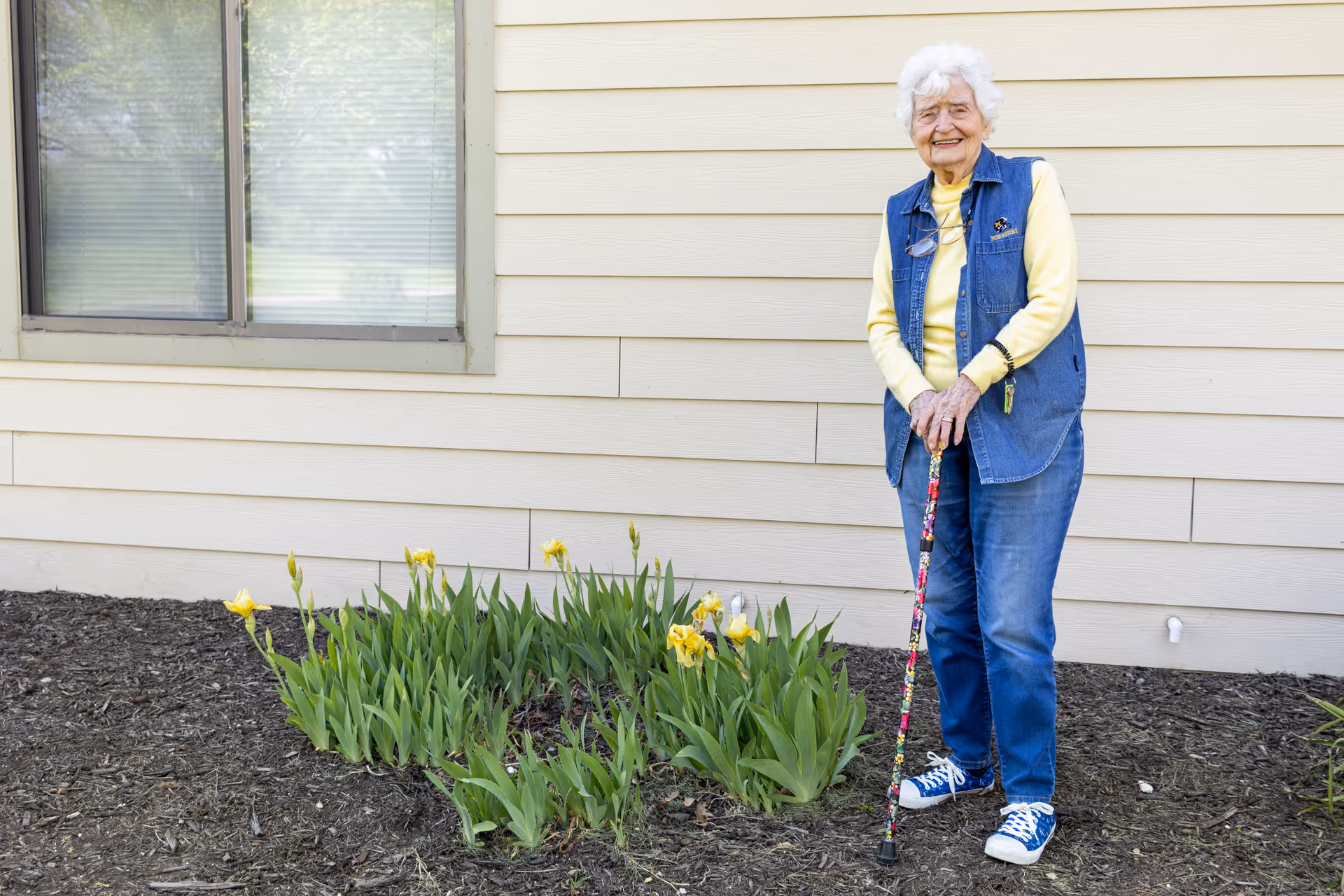 An elderly woman with white hair standing outside next to a flower bed with yellow flowers. She is smiling, wearing a yellow long-sleeve shirt, a blue denim vest, blue jeans, and blue sneakers. She is holding a colorful cane and standing in front of a beige building with a window.