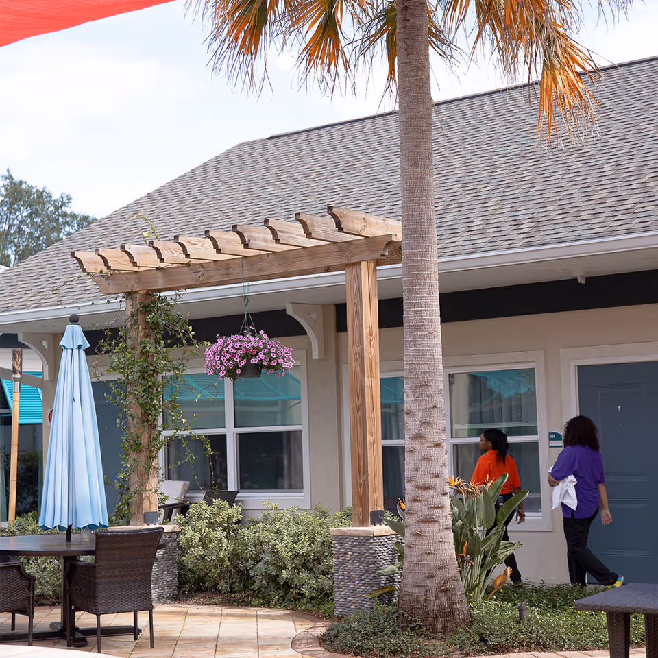Outdoor courtyard of an assisted living facility with a wooden pergola, palm tree, patio furniture, hanging flowers, and two people walking by.