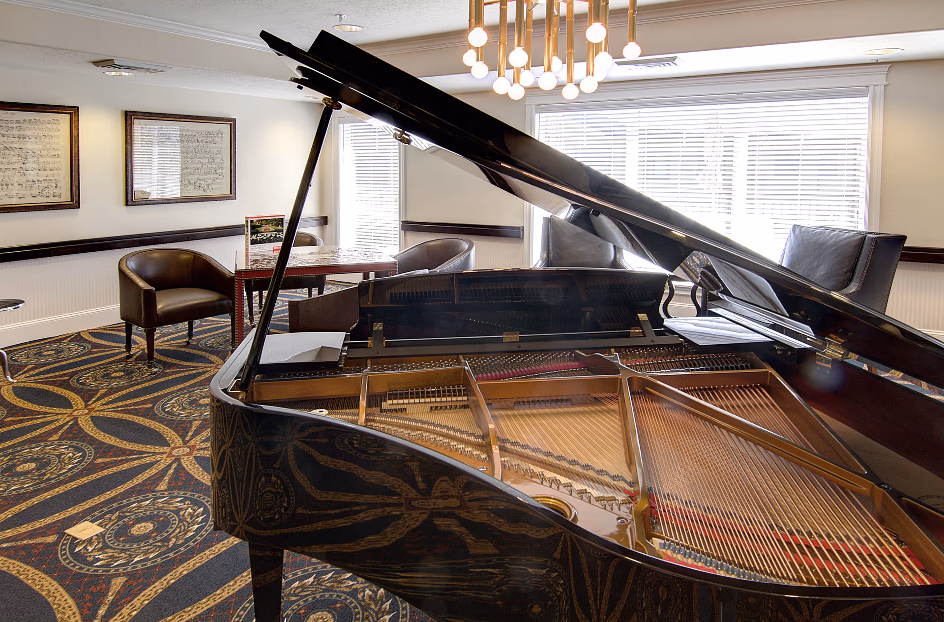 Interior room with a grand piano in the foreground, a patterned carpet, two leather chairs around a small table, framed sheet music on the wall, and large windows with blinds letting in natural light.