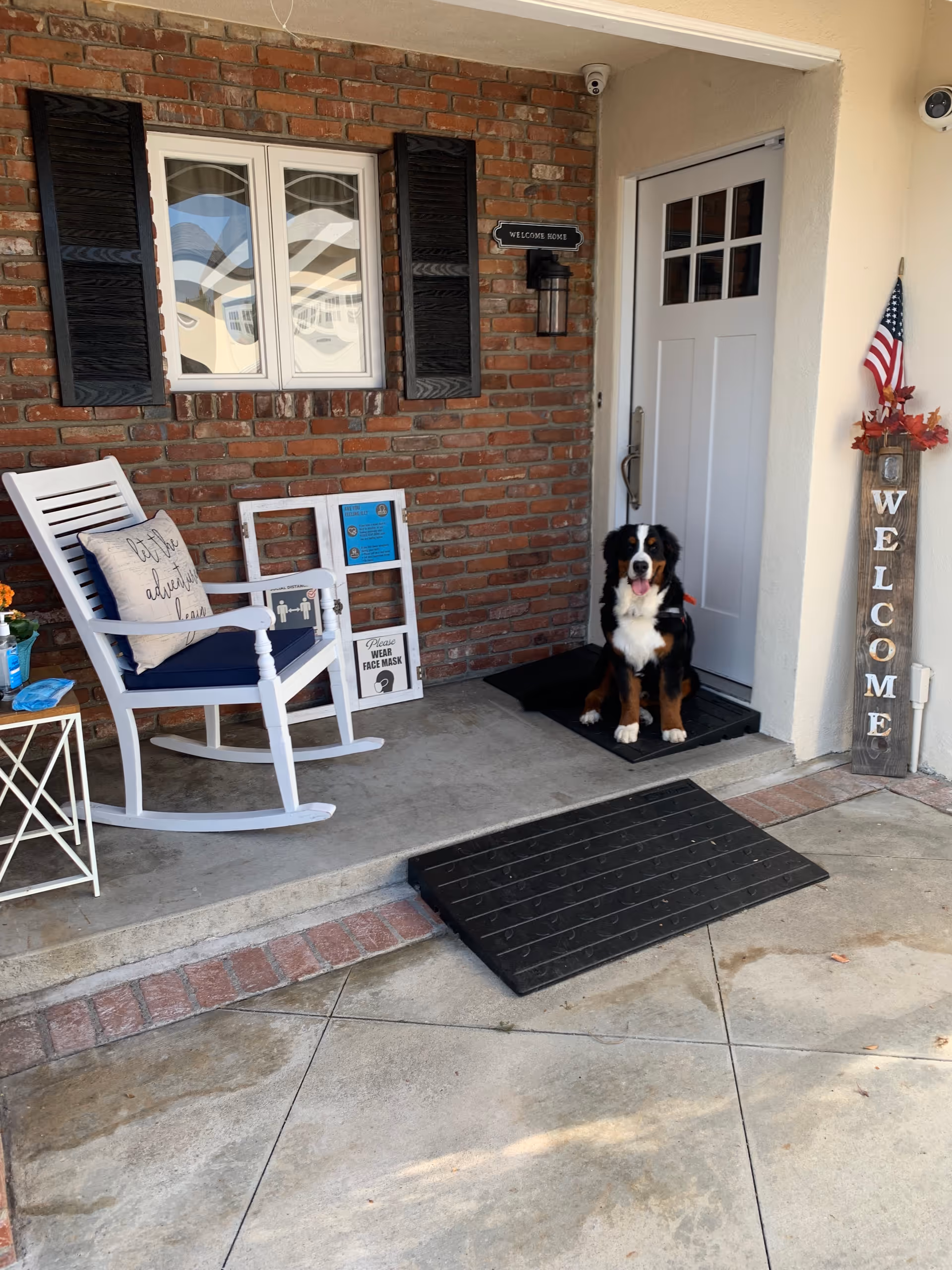 A porch area with a white rocking chair that has a cushion and a pillow with the text 'Let the adventure begin'. There is a small white side table with a blue bottle and some items on it. A black and white dog is sitting on a black mat in front of a white door with glass panes. The brick wall behind the chair has a window with black shutters and a sign that says 'WELCOME HOME'. Next to the door is a vertical wooden sign with the word 'WELCOME' and an American flag with fall decorations on top.