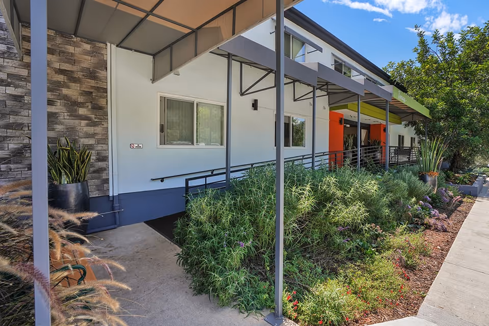 Exterior view of a senior living facility entrance with a covered walkway, access ramp, and landscaped garden beds.