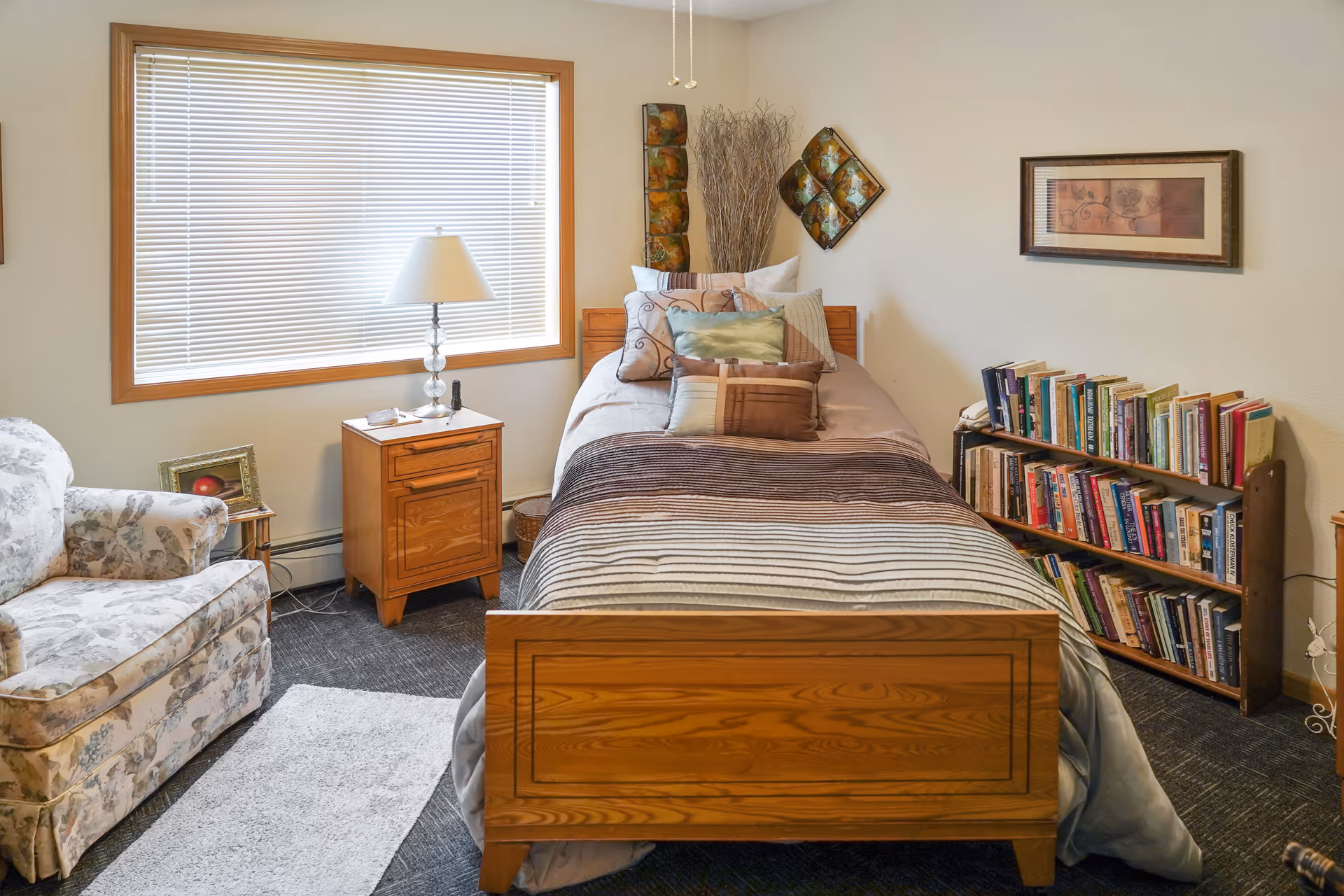 A cozy single bedroom with a wooden bed, bedside table and lamp, upholstered chair, and a bookshelf filled with books.