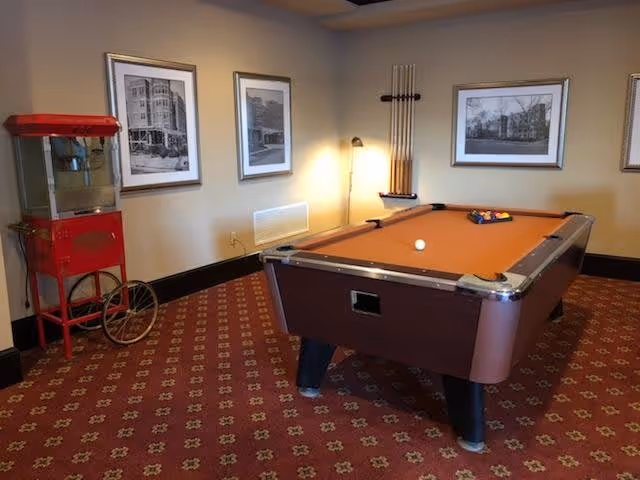 A carpeted game room with an orange-felt pool table, cue rack, framed black-and-white photos on the walls, a floor lamp, and a red popcorn machine.