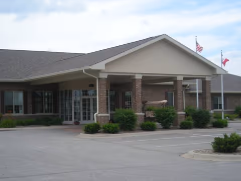 Exterior view of a single-story senior living facility building with a covered entrance supported by brick columns. There are two flagpoles with flags flying near the entrance and some shrubs and greenery around the building. The parking lot is visible in the foreground.