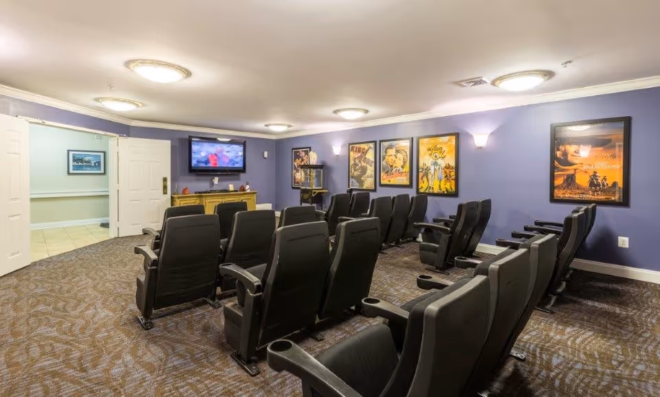 A small movie theater room with two rows of black cushioned seats facing a wall-mounted flat screen TV. The walls are painted purple and decorated with framed vintage movie posters. The room has carpeted flooring and ceiling lights. Double doors lead to an adjacent hallway.