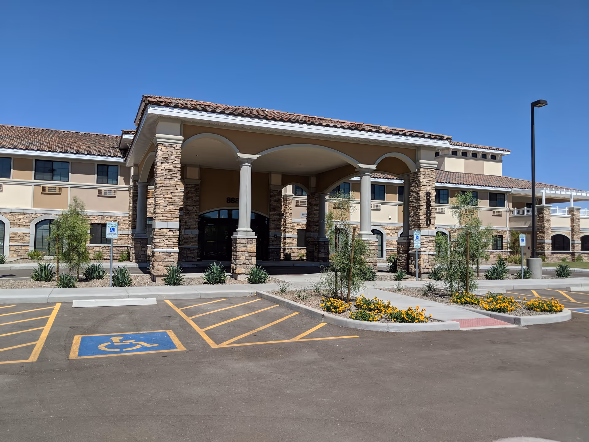 Exterior view of The Mission at Agua Fria Senior Living building with stone and stucco facade, arched entrance supported by columns, handicap parking spaces, and landscaped areas with small trees and yellow flowers under a clear blue sky.