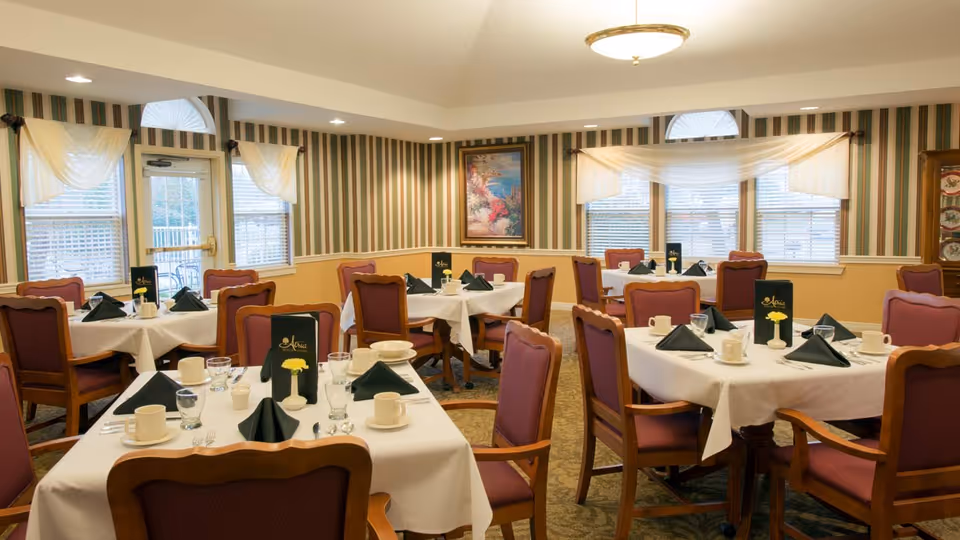 A dining room with multiple tables covered in white tablecloths, each set with cups, glasses, plates, silverware, black folded napkins, and small yellow flower centerpieces. The room has striped wallpaper, several windows with white curtains, and framed artwork on the walls. Wooden chairs with purple cushions surround the tables.
