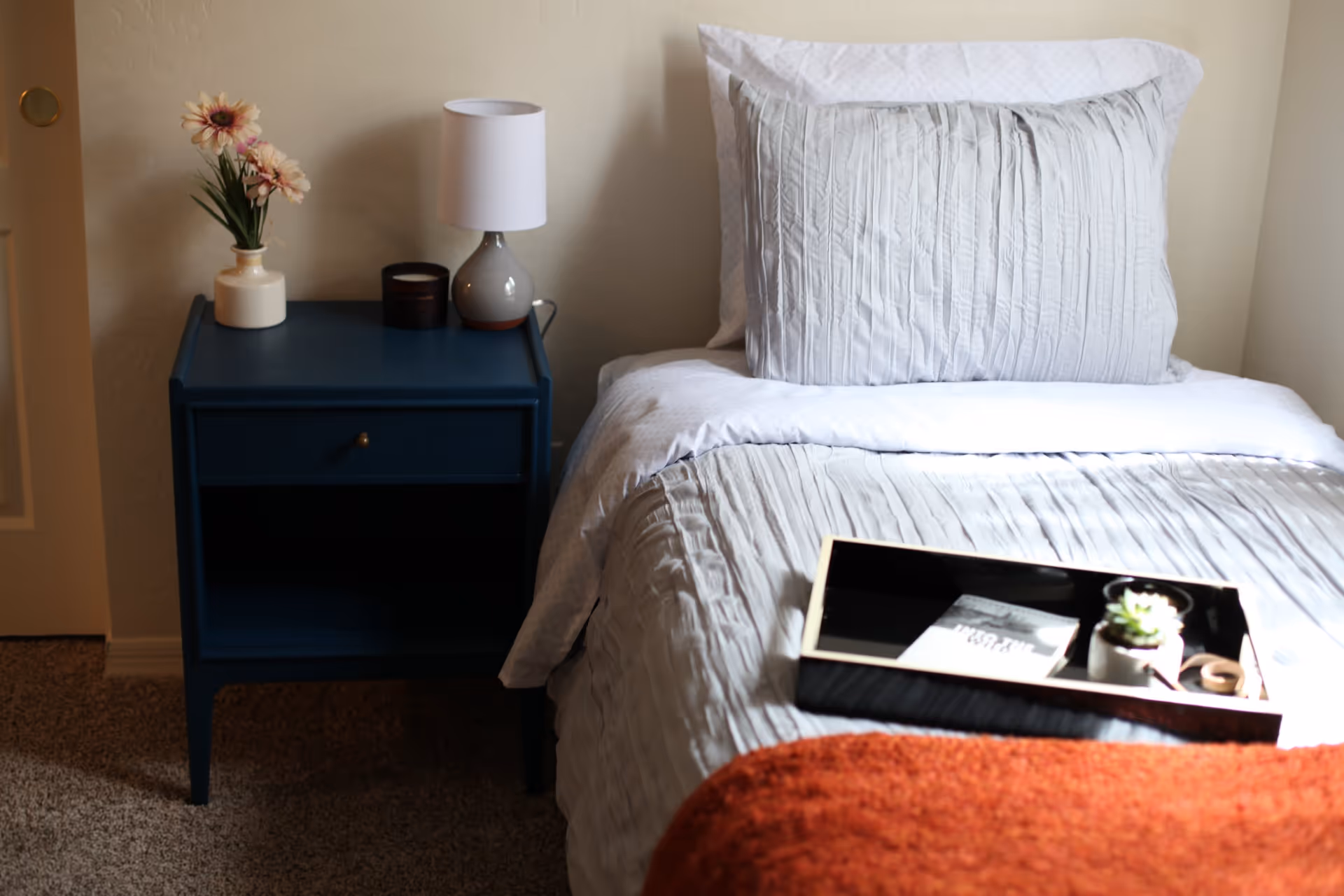 A neatly made bed with white and light gray bedding, a large pillow, and an orange blanket at the foot. On the bed is a black tray holding a small potted succulent and a book. Next to the bed is a dark blue nightstand with a drawer, holding a small lamp, a candle, and a vase with light pink flowers. The room has beige walls and carpeted floor.