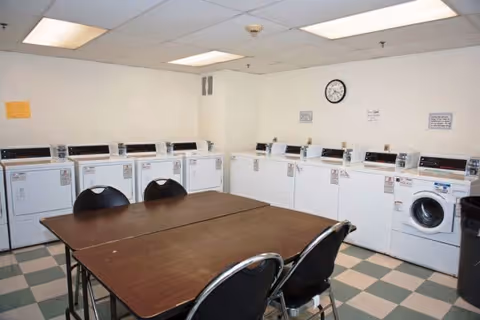 Communal laundry room with washers and dryers lined along the walls, a central table with chairs, and a wall clock.