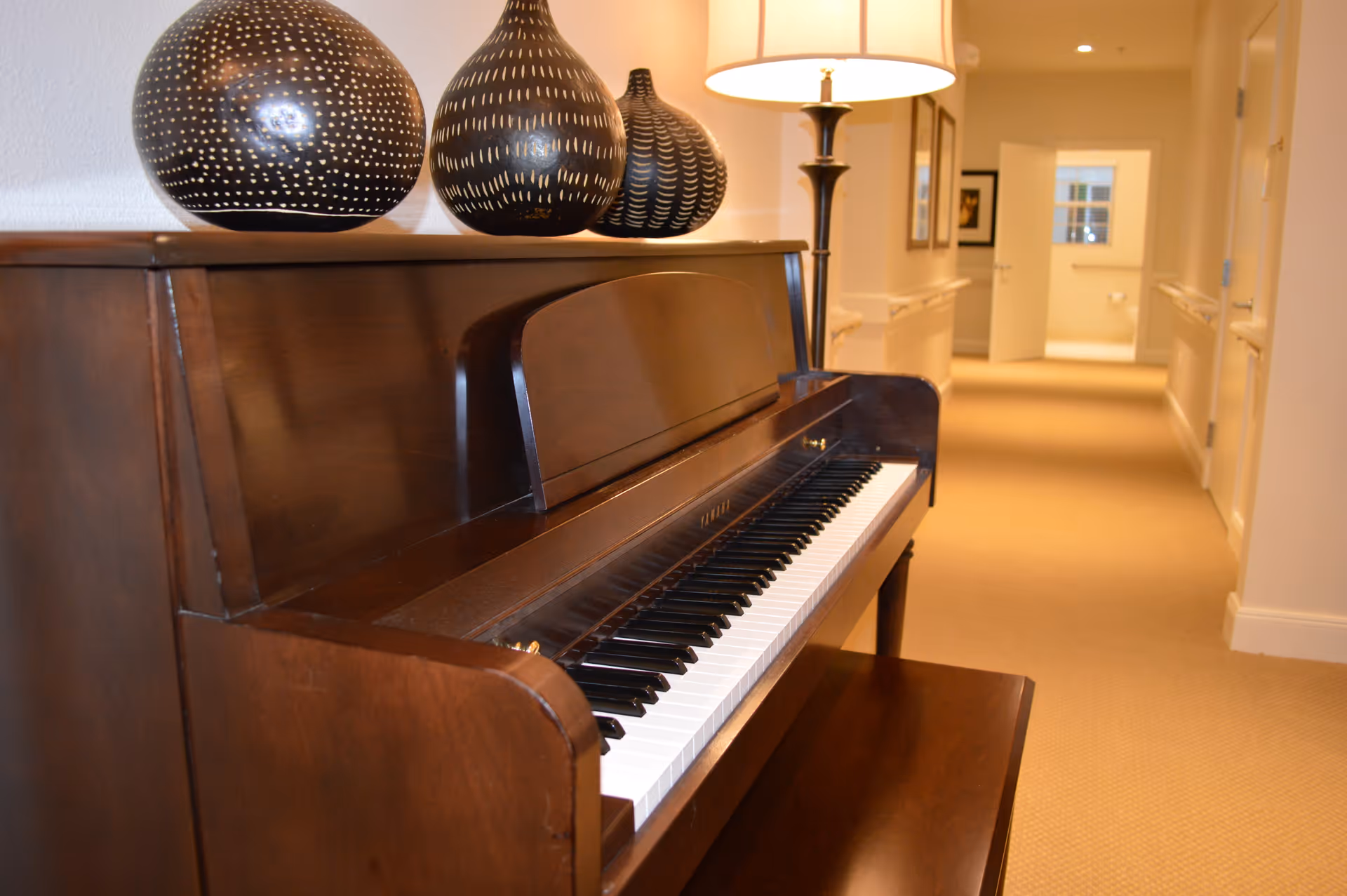 A wooden upright piano with three decorative vases on top and a lamp beside it, placed in a well-lit hallway with beige carpet and cream-colored walls. The hallway has handrails on both sides and doors leading to other rooms.
