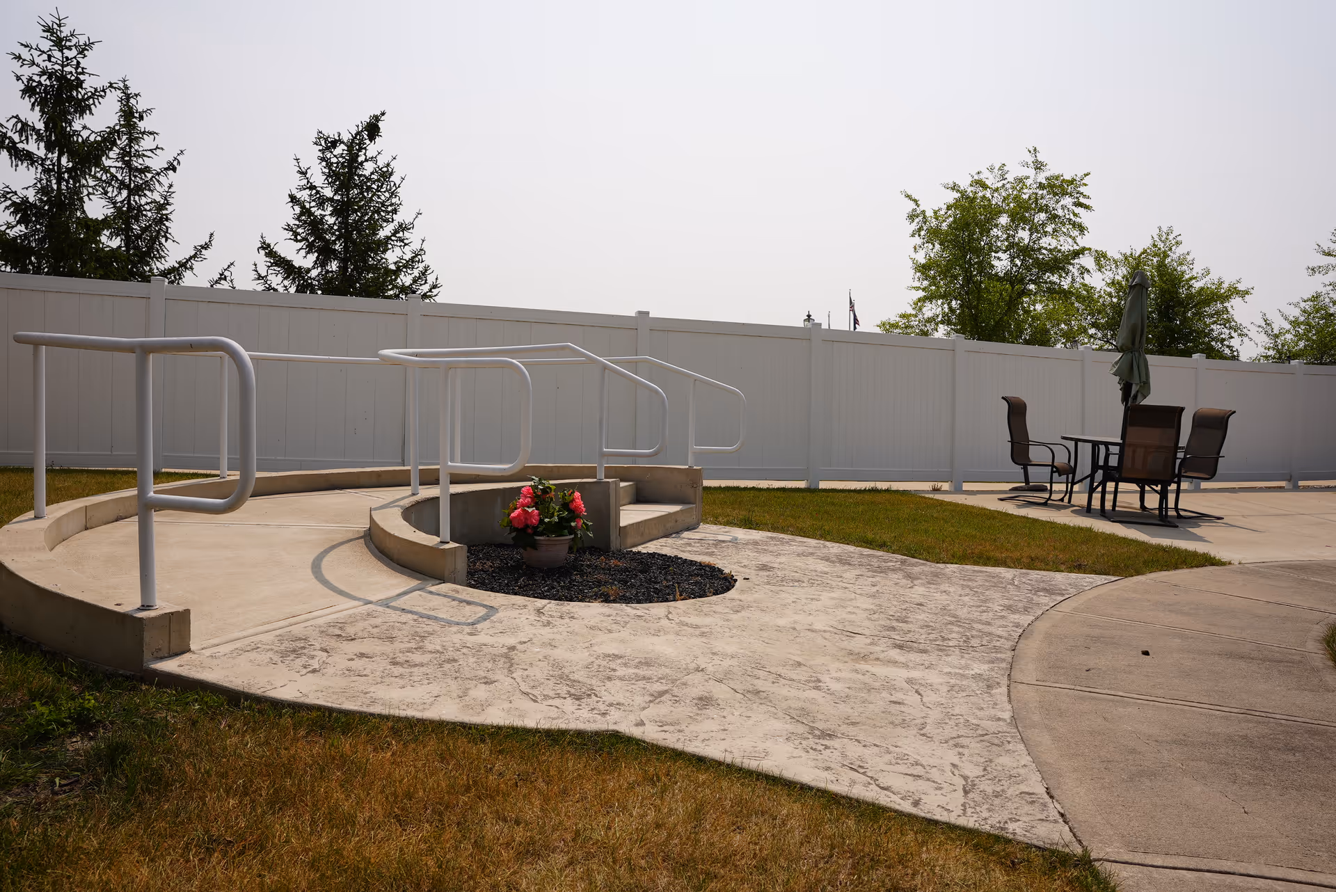 Outdoor patio area with a curved concrete ramp featuring white handrails, a small flower pot with pink flowers in the center, a white privacy fence in the background, and a table with four chairs and a closed umbrella on a concrete surface to the right.