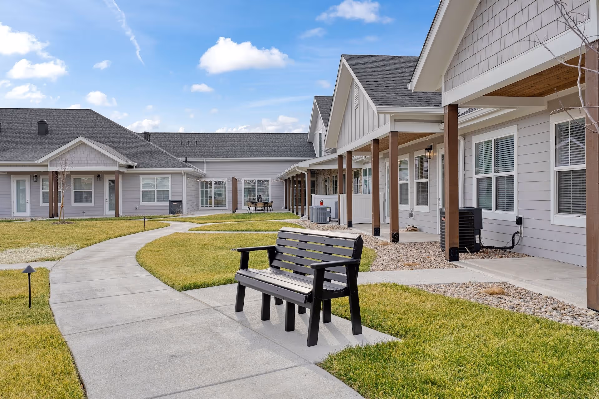 Courtyard with a curved concrete walkway and a bench in front of single-story senior living residences under a blue sky.