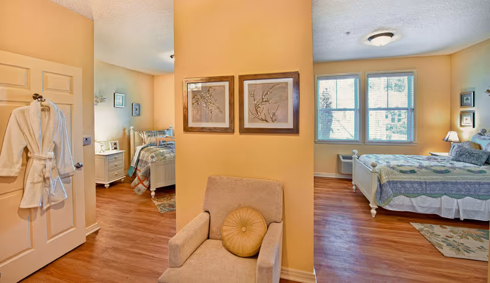 Interior view of a senior living facility room with two bedrooms visible. Each bedroom has a bed with patterned bedding, nightstands with lamps, and framed artwork on the walls. A beige armchair with a round cushion is positioned in the foreground against a yellow wall. A white bathrobe hangs on a door to the left. The rooms have wooden floors and large windows letting in natural light.