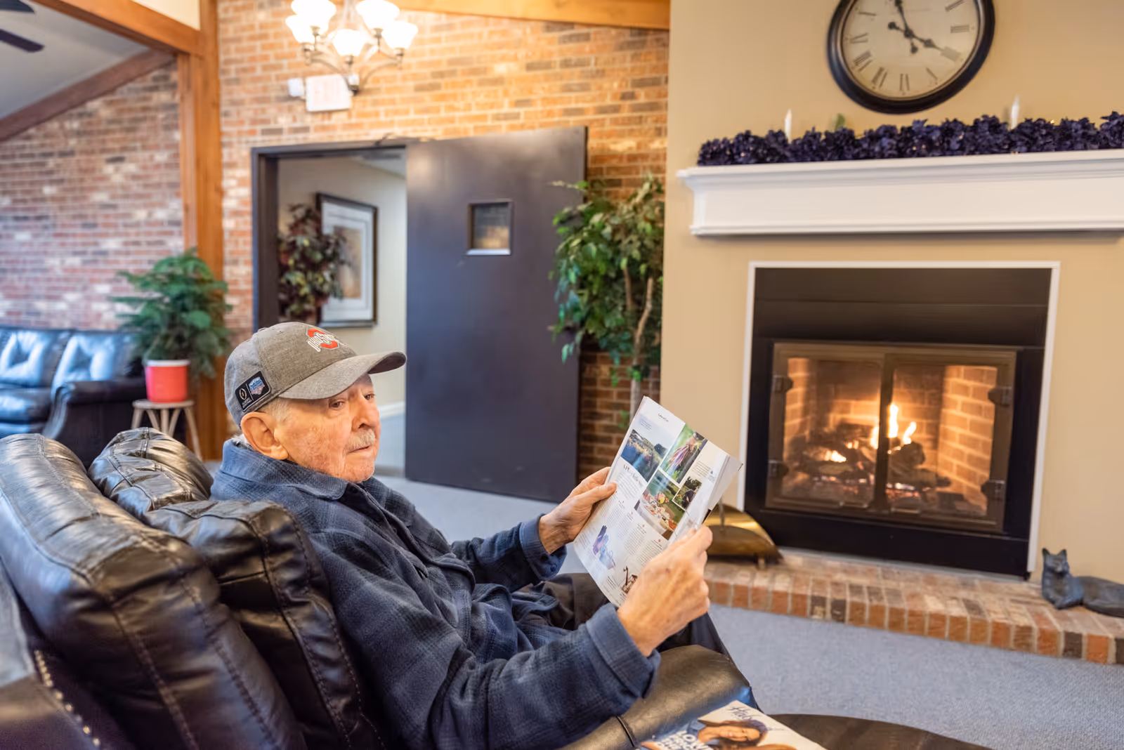 An elderly man wearing a gray cap and blue jacket is sitting on a black leather couch in a cozy room with a lit fireplace. He is reading a magazine. The room has brick walls, a clock above the fireplace, plants, and a door in the background.