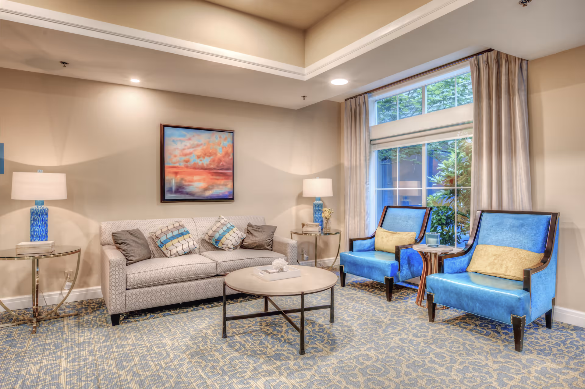 A cozy living room area with a beige sofa adorned with four decorative pillows, two blue armchairs with yellow cushions, a round coffee table in the center, two side tables each with a blue lamp, a large window with beige curtains, and a colorful painting of a sunset on the wall.