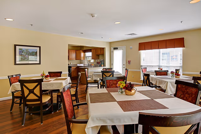 A bright dining room with several tables covered in white tablecloths and brown placemats, each decorated with small flower arrangements. The room has wooden chairs with red patterned cushions, hardwood floors, beige walls, and a large window with blinds letting in natural light. In the background, there is a kitchen area with wooden cabinets and a door leading outside.