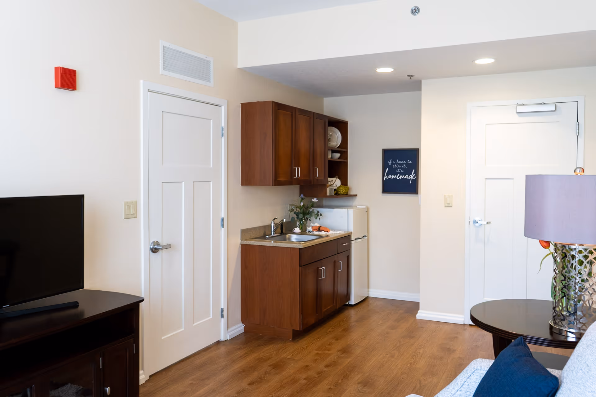 Interior view of a small kitchen area in an assisted living facility with wooden cabinets, a countertop with a sink, a small white refrigerator, and a decorative sign on the wall. To the left, there is a TV on a dark wooden stand, and to the right, part of a round table with a lamp and a couch with a blue pillow are visible. The floor is wooden, and there are two white doors in the background.