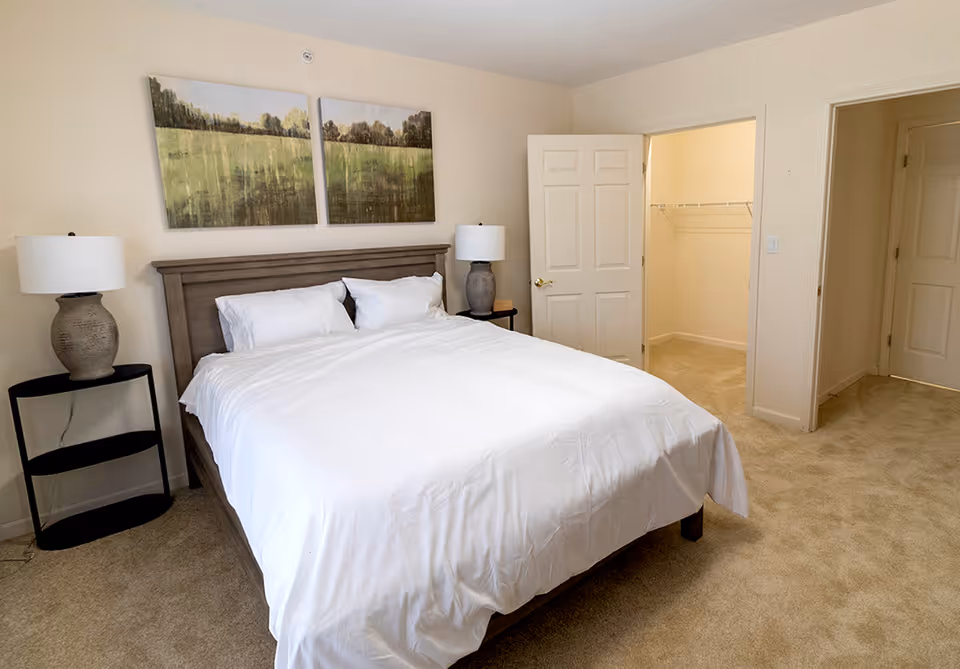 A bedroom with a large bed covered in white bedding and pillows. There are two bedside tables, each with a lamp. Above the bed are two landscape paintings depicting a green field and trees. The room has beige carpet and light-colored walls. There are two open doors, one leading to a walk-in closet and the other to another room or hallway.