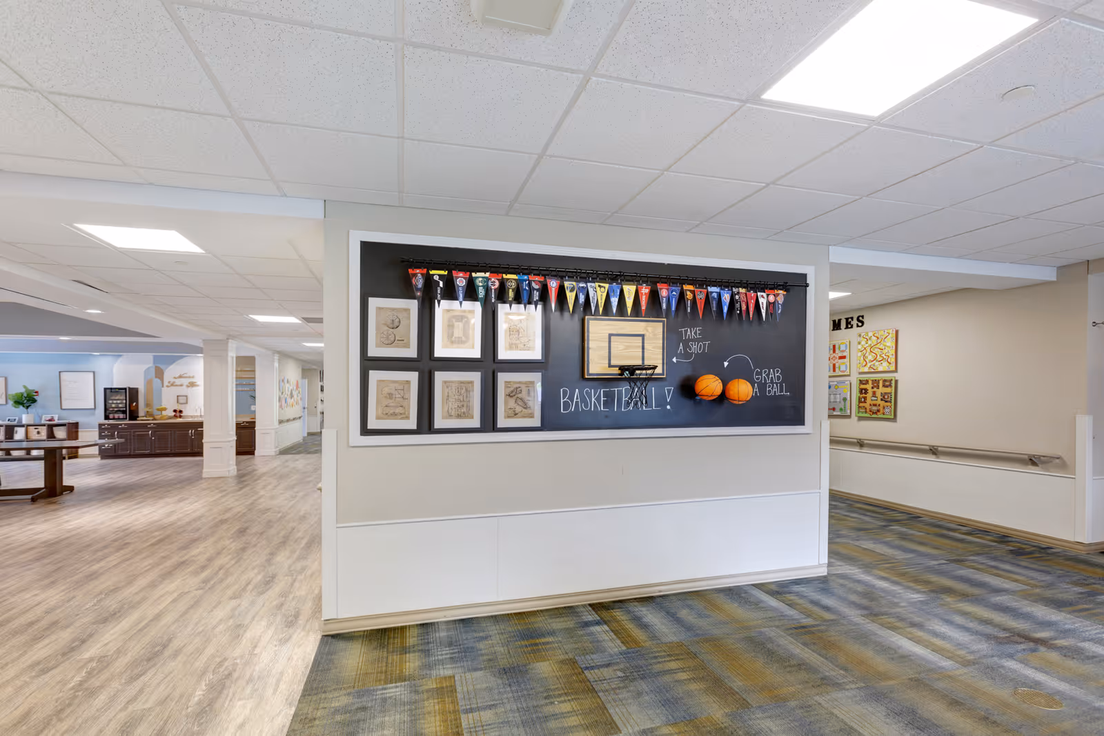 Interior hallway of a senior living facility with a basketball-themed bulletin board displaying small pennants, framed pictures, and two basketballs. The hallway has a mix of wood and carpet flooring, with handrails along the walls and a seating area visible in the background.