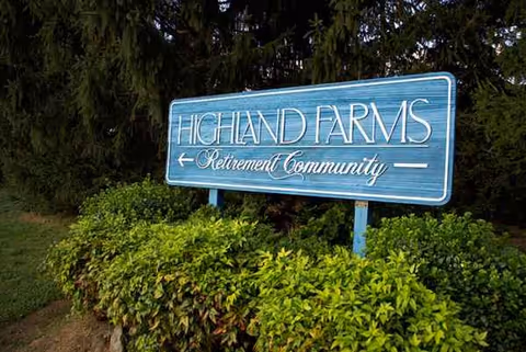 Blue Highland Farms Retirement Community sign with green shrubs in foreground
