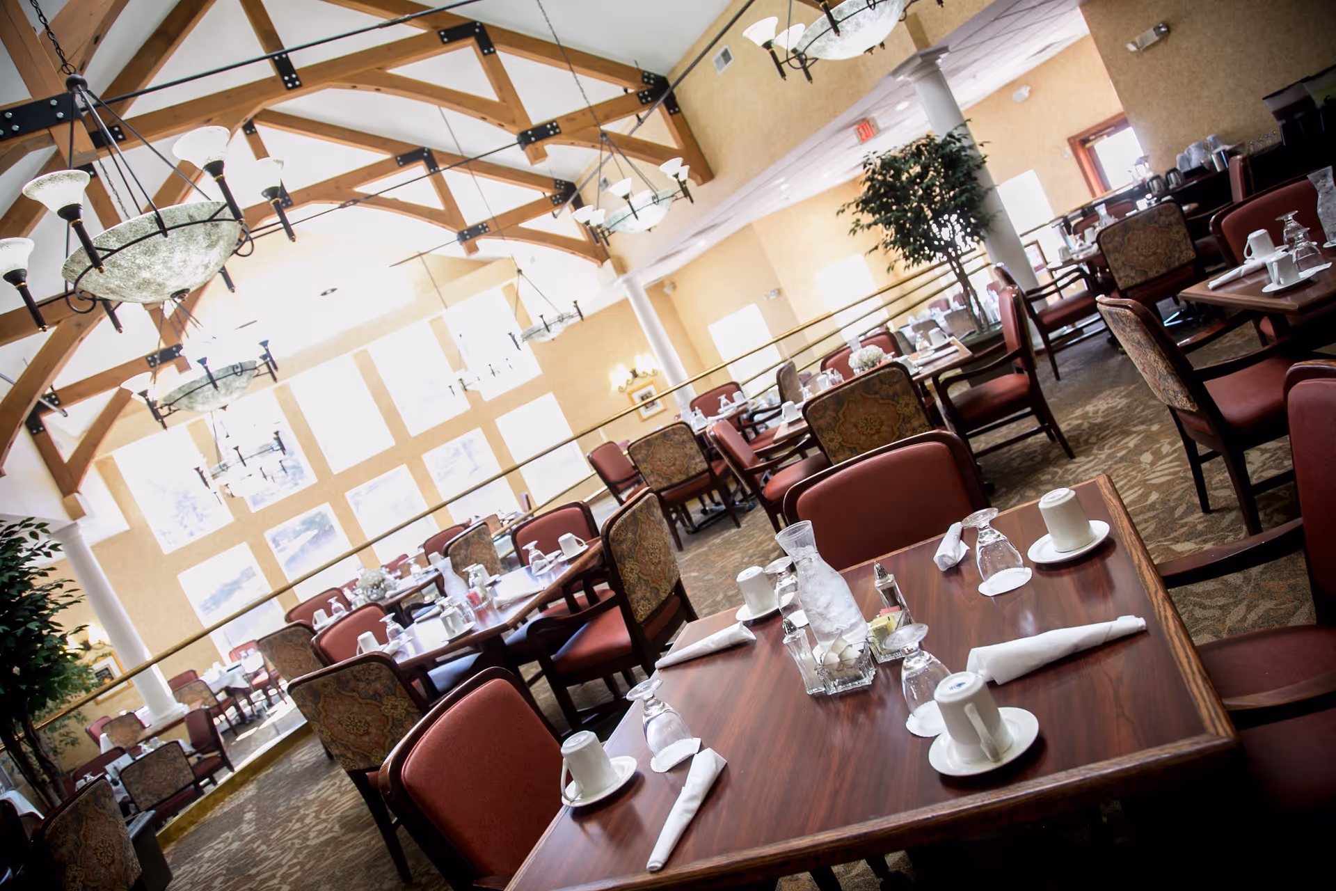 A spacious dining room with multiple tables set with white napkins, cups, glasses, and a water pitcher. The room features large windows allowing natural light to fill the space, high ceilings with wooden beams, and elegant hanging light fixtures. Upholstered chairs with patterned backs and red seats surround the tables, and there are some potted plants adding greenery to the room.