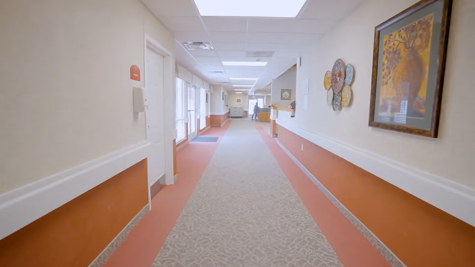A long, well-lit hallway in an assisted living facility with beige walls and a patterned carpet. The lower half of the walls is painted orange with white trim. There are framed artworks and decorative plates on the right wall. At the end of the hallway, a person is visible near a reception or service desk area.