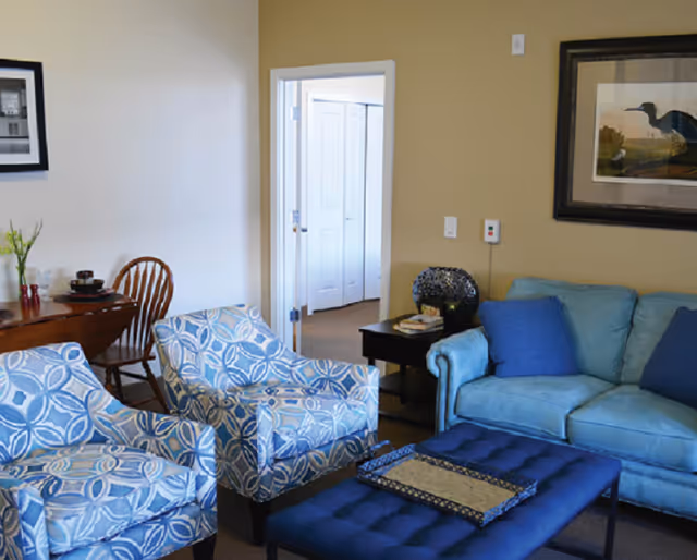 A cozy living room with a light blue sofa adorned with blue cushions, two patterned blue armchairs, a dark blue ottoman with a decorative tray, a small black side table with books and a lamp, and a wooden dining table with chairs in the background. A framed bird artwork hangs on the beige wall.