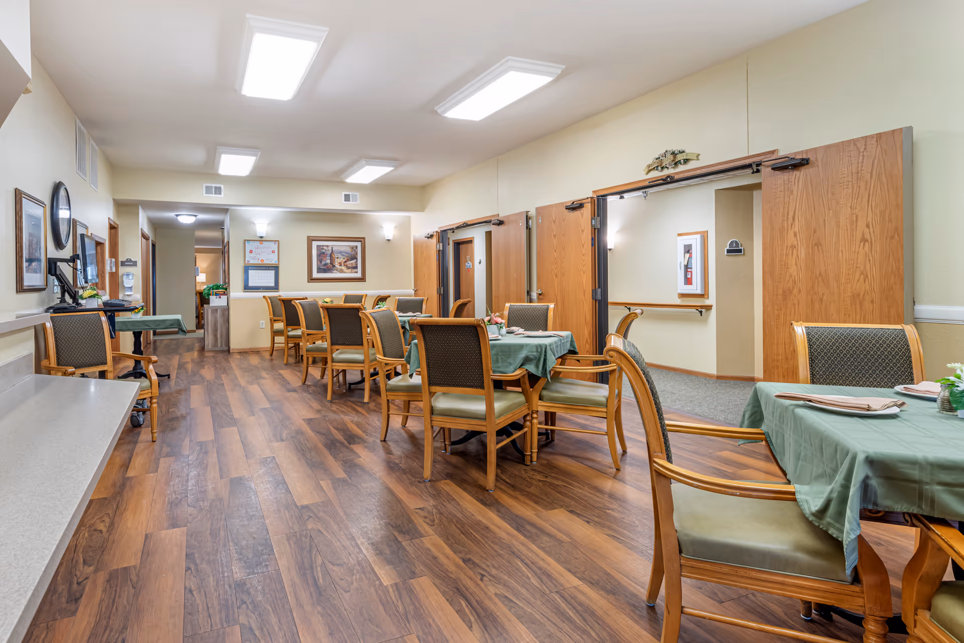 Dining room with tables covered in green tablecloths, wooden chairs, and wood-look flooring in a senior living facility.