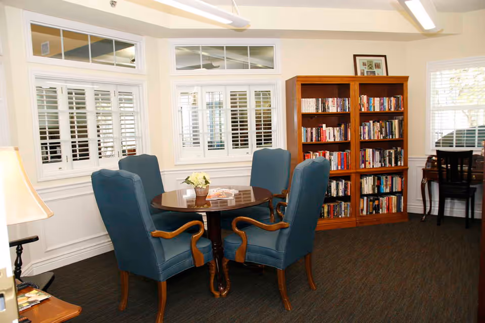A cozy room with a round wooden table surrounded by four blue upholstered chairs with wooden arms. Behind the table is a wooden bookshelf filled with books. The room has white walls with large windows featuring white shutters, allowing natural light to enter. A small desk with a chair is positioned near one of the windows, and a lamp is visible on the left side.