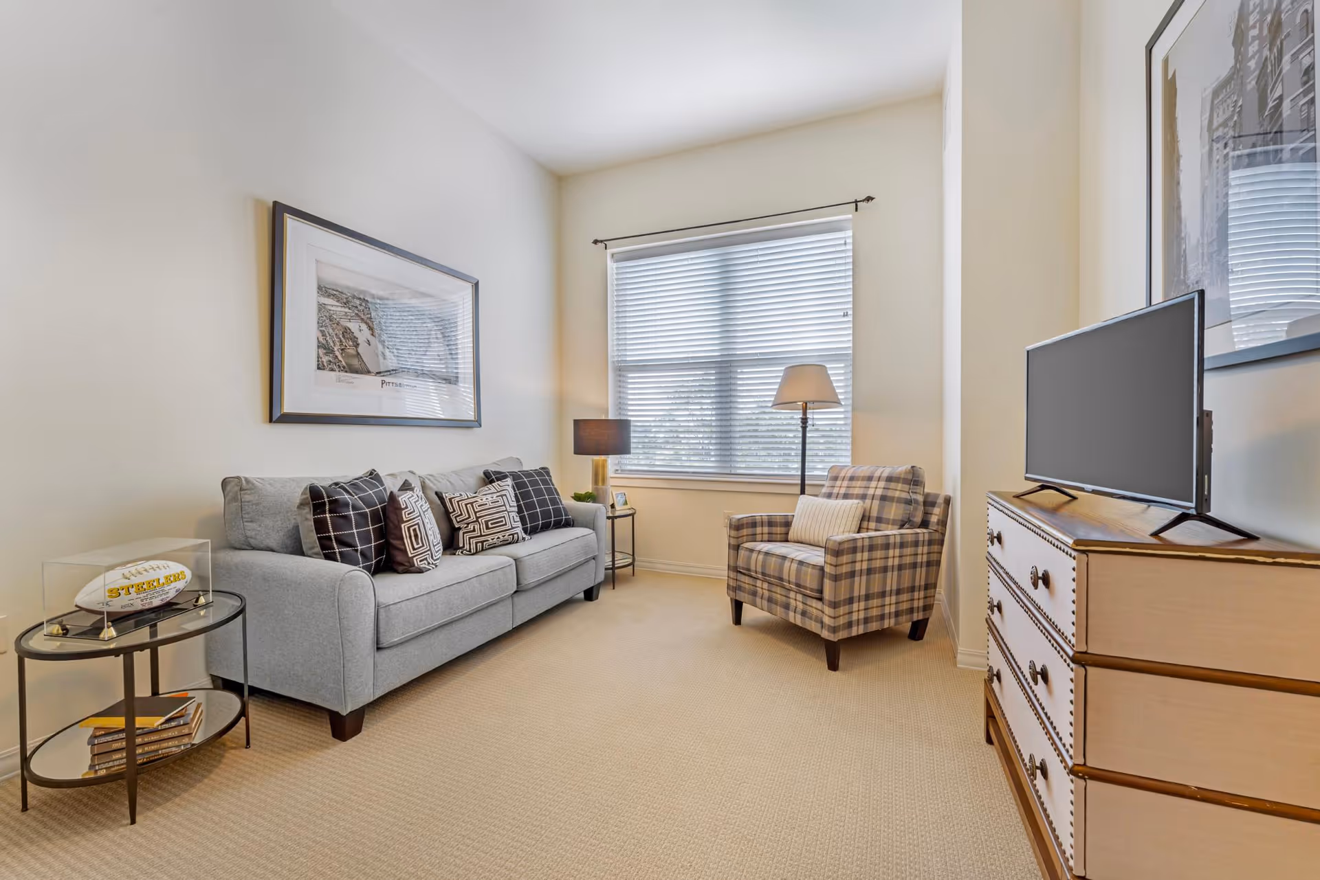 Bright living room with a gray sofa, patterned armchair, window, TV on a dresser, and side tables with lamps.