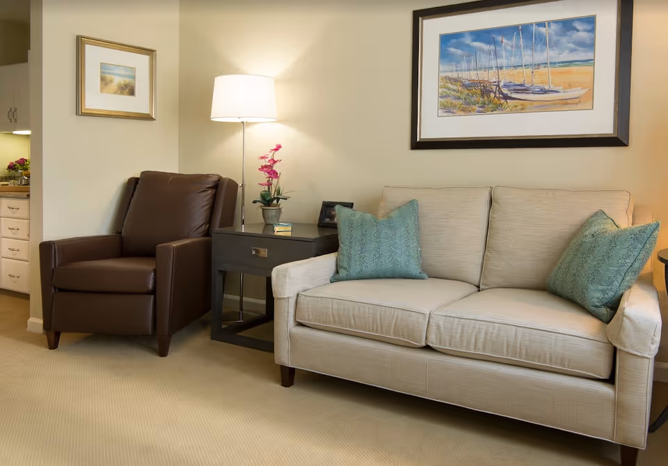 A cozy living room area featuring a beige sofa with two green patterned pillows, a dark brown leather armchair, a black side table with a potted pink orchid and books, a floor lamp, and two framed paintings on the walls.