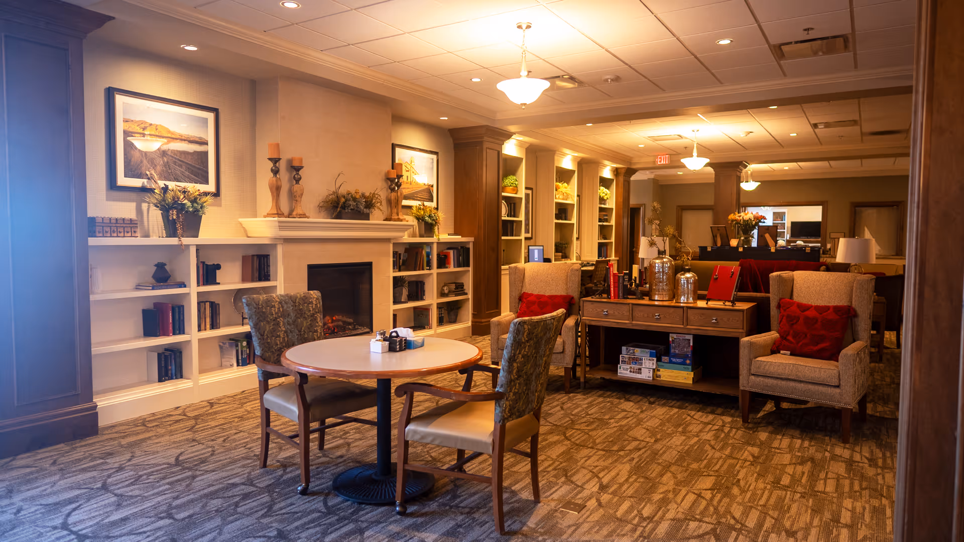 A cozy senior living common area with a round table and four chairs in the foreground. The room features a fireplace with decorative candles and plants on the mantel, built-in bookshelves filled with books and decor, and comfortable armchairs with red cushions. Warm lighting and carpeted floors create a welcoming atmosphere.