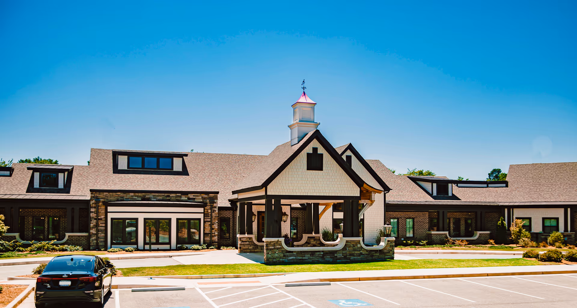 Exterior view of a single-story building with a peaked roof and a small tower-like structure on top. The building has large windows and a covered entrance with stone and wood accents. A black car is parked in the parking lot in front of the building under a clear blue sky.