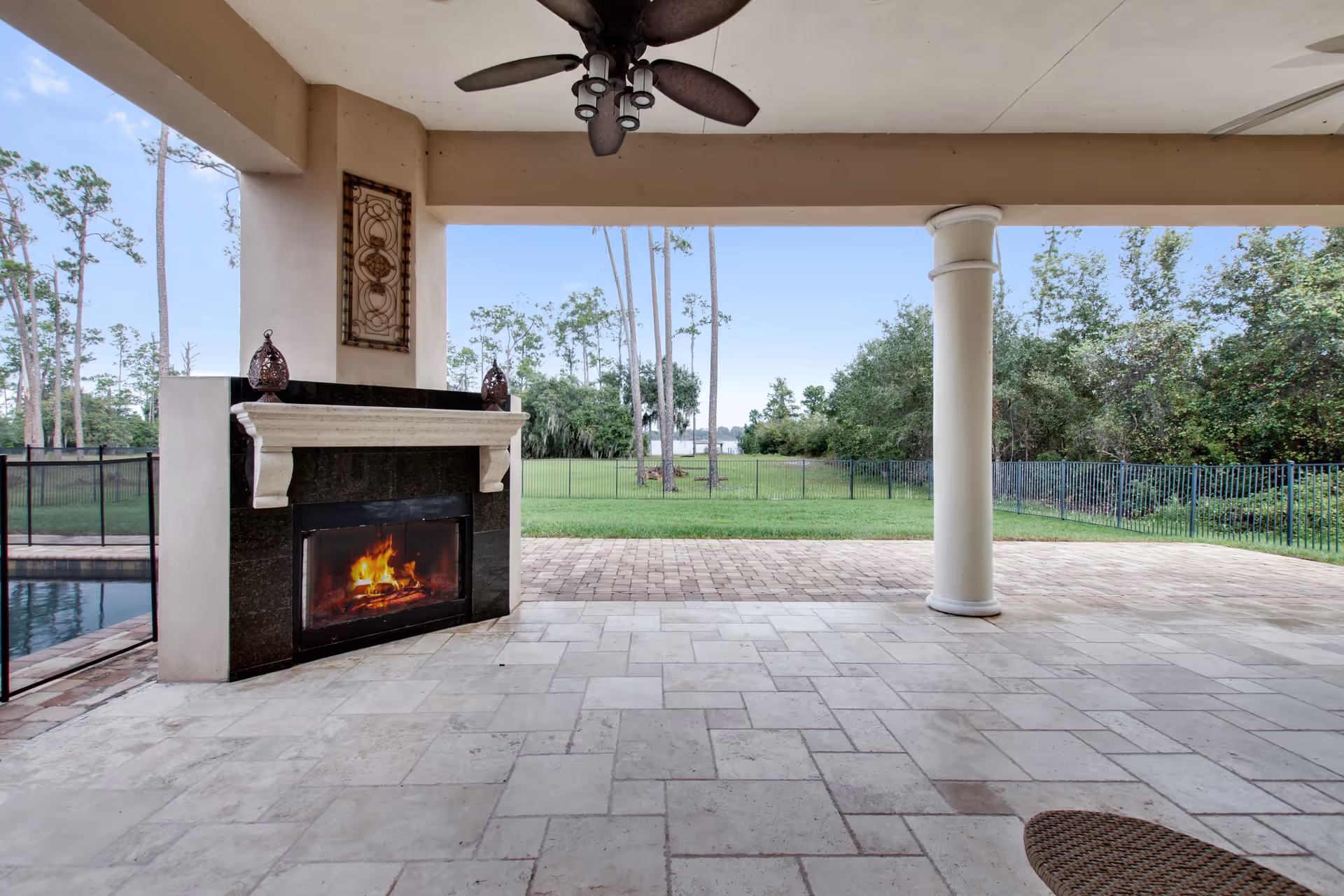 Covered outdoor patio area with a ceiling fan, a lit fireplace with a black and white mantel, tiled flooring, and a view of a grassy yard with trees and a fence in the background.