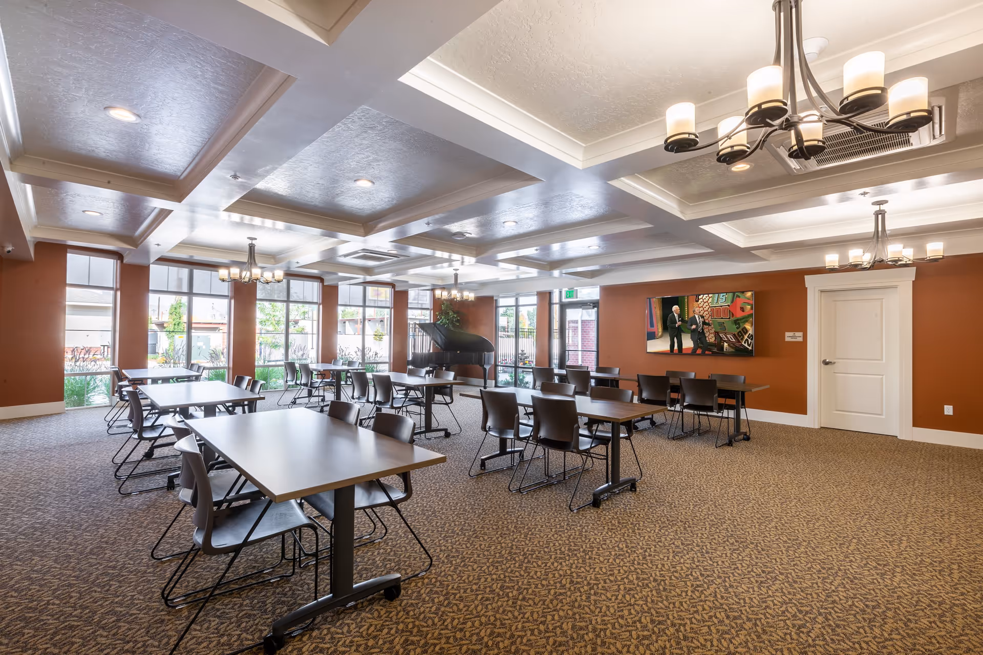 Spacious multipurpose dining room with rows of tables and chairs, chandeliers, large windows, and a piano near the back.